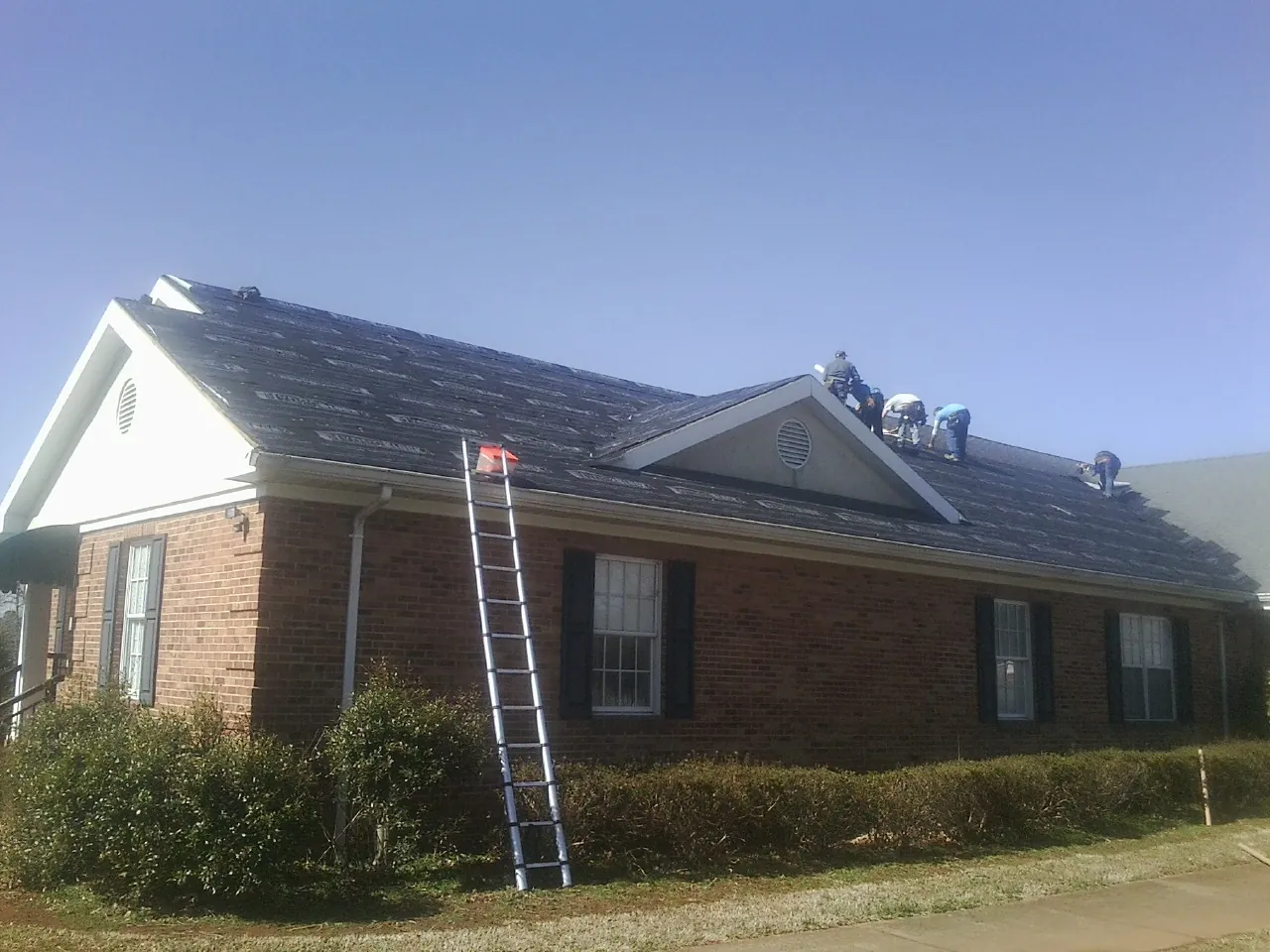 Crew tearing shingles off a brick ranch roof with a ladder set — Metal Roof Repair work by our crew in Corinne
