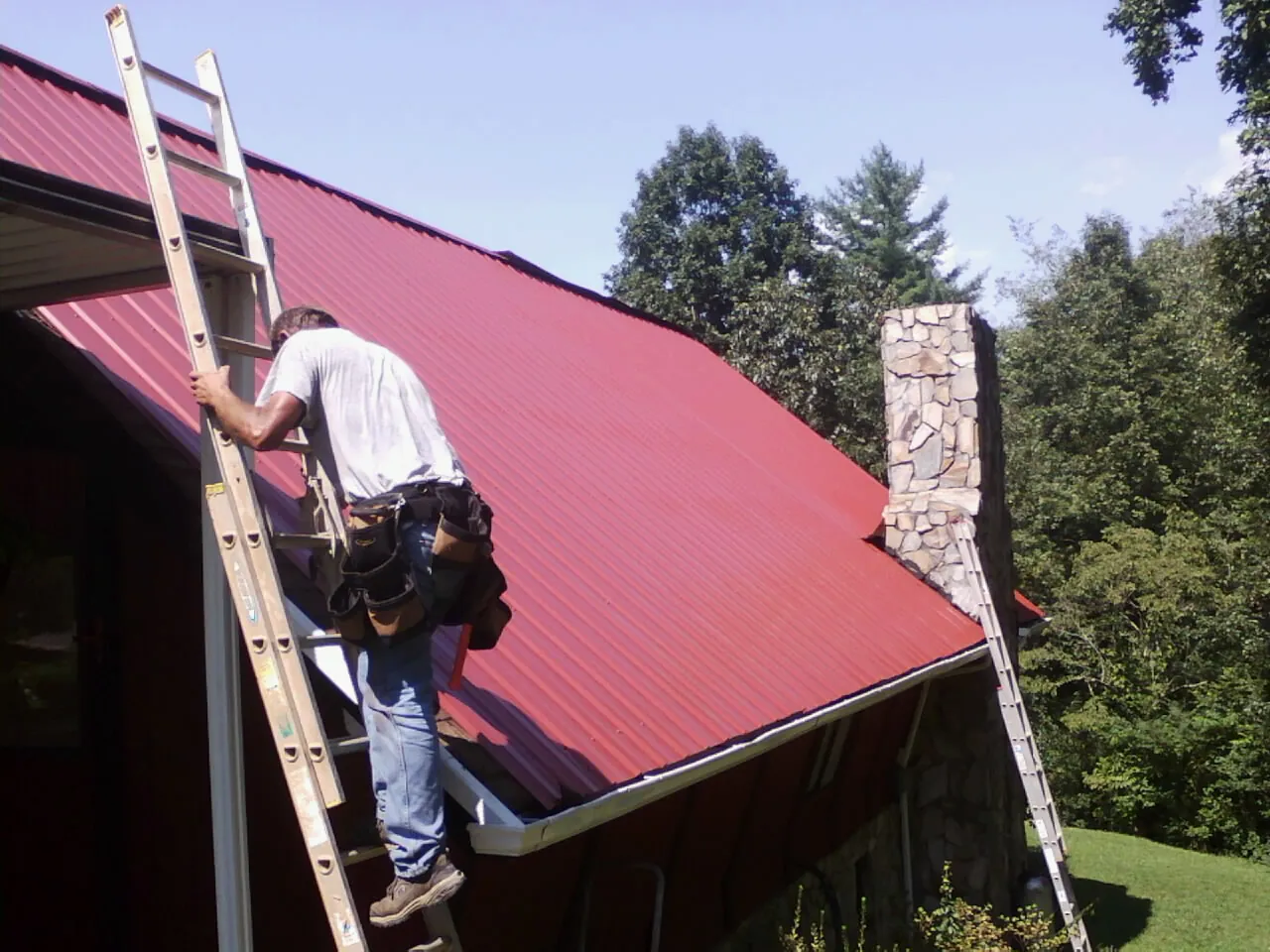 Roofer on a ladder beside a red metal roof and stone chimney — Soffit & Fascia Repair by Roofing Brothers in Corinne